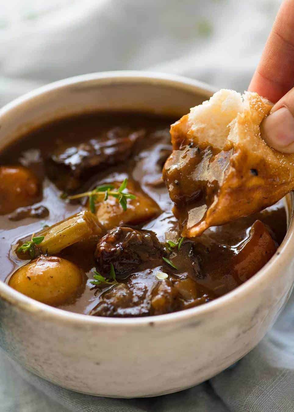 Close up photo of a piece of bread being dunked into Beef Stew with Potatoes and Carrots. The bread shows how thick the gravy sauce is./